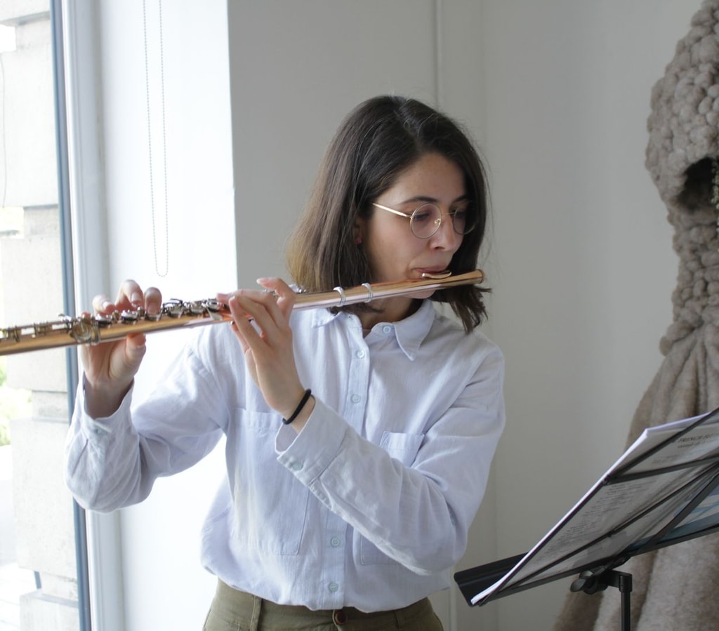 Woman with dark hair and wearing a blue button-up shirt plays the flute, whilst a sculpture looking like a hooded figure looms behind her.