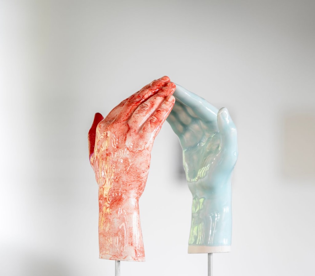 Two ceramic hands, one glazed in red and one in pale blue, suspended on metal rods. Their fingertips almost touch. By Maud Haya-Baviera.