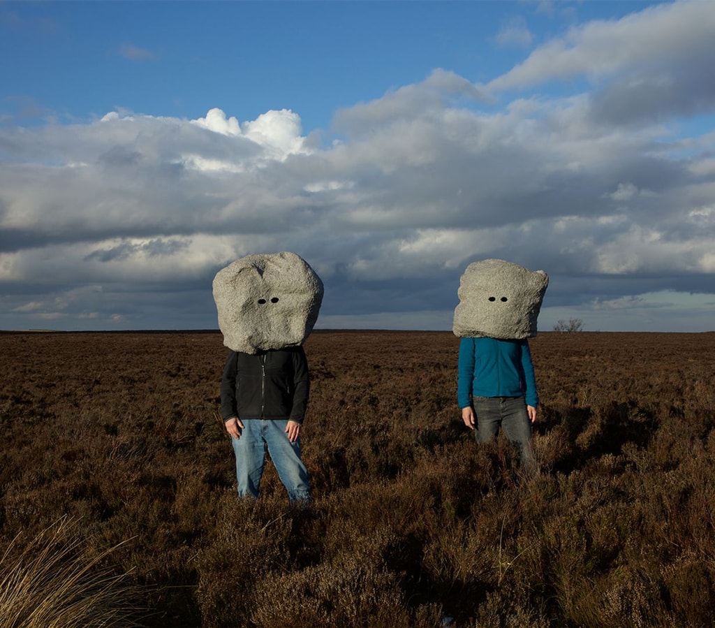 Photograph by Conroy / Sanderson, two people standing in a remote grassland. Their heads are covered by large boulders with eye holes.