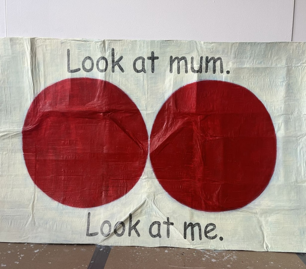 Crumped and creased piece of cardboard, painted pale yellow with two dark red circles in the centre, almost touching. Text reads "Look at mum. Look at me."