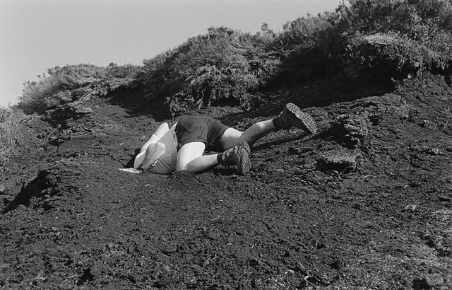 Black and white photograph by David Eckersley, man in hiking gear on a hilltop with his head buried in the ground.