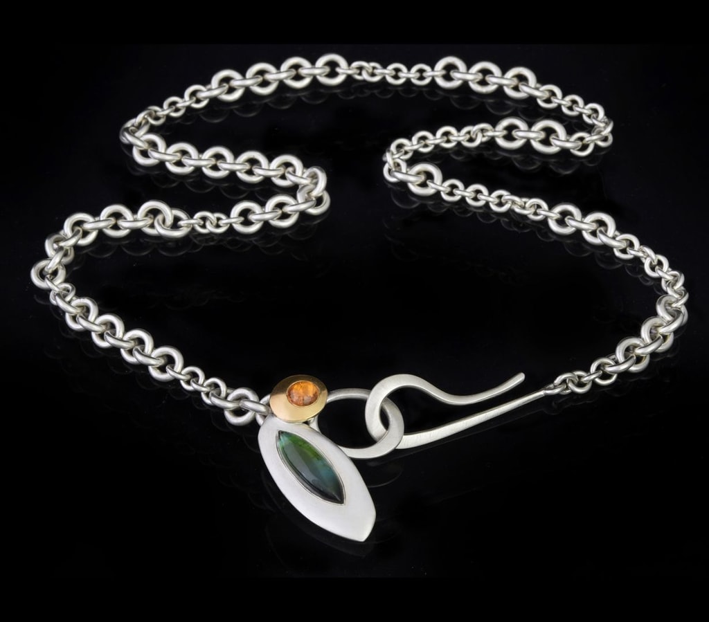 Necklace by Emily Thatcher, chain with varied thickness and a large hook fastener, with silver and gold pendants featuring green and orange gemstones.
