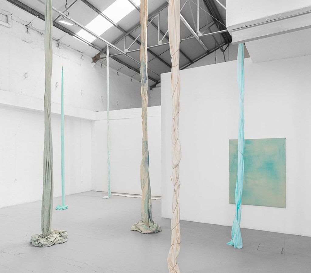 Installation by Jo Fleming Smith in a tall white gallery, lonf pieces of twisted blue fabric hang from the ceiling and bunch on the floor. Blue canvas in the background.