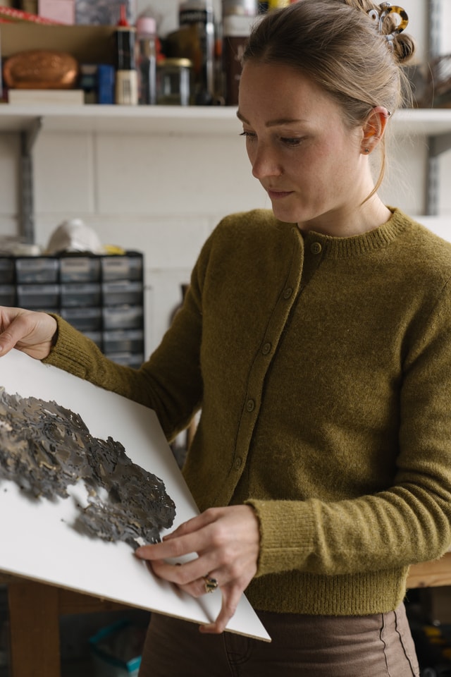 A young white woman in an olive green jumper stands in her studio holding some work she has made, an intricate cut-out piece of metal.