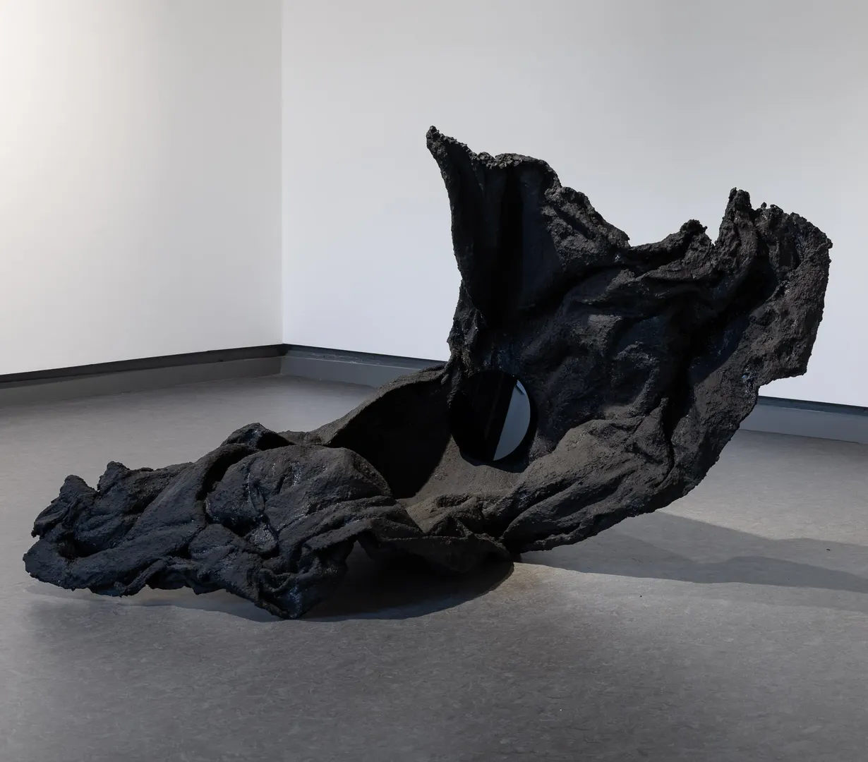 Black fabric hardened into a solid form that lifts off the ground, looks like stone or part of a cliffside. A small round black mirror sits within a crevice.